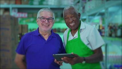 Joyful senior diverse colleagues of supermarket smiling at camera. Two older staff managers of supermarket standing at aisle