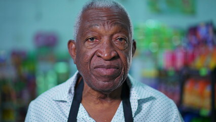 One senior black male employee of supermarket standing inside supermarket wearing apron with neutral expression. Portrait of African American person staff of grocery store