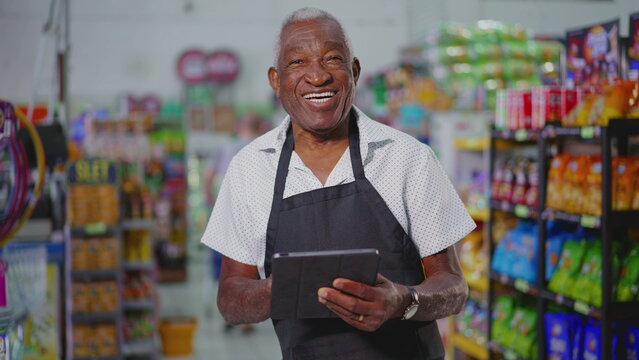 Joyful Happy Brazilian senior employee of supermarkert wearing apron and holding tablet device, portrait of a black older male staff depicting job occupation at retail store