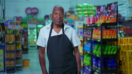 One senior black male employee of supermarket standing inside supermarket wearing apron with neutral expression. Portrait of African American person staff of grocery store