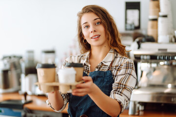 Portrait of a young woman barista holding takeaway coffee in her hands. Owner of the coffee shop stands behind the bar in an apron and gives coffee to go. Takeaway food. Business concept.