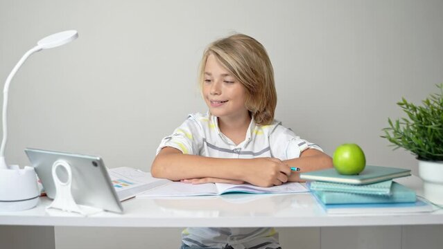 Middle school smiling student boy sitting at desk, remote studying writing book homework and tablet at class home e-learning Back to school. Childhood, education for children