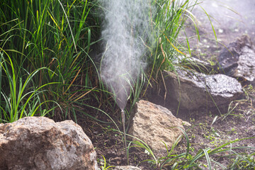 The nozzle of the sprinkler system sprays water into small droplets to irrigate plants in a landscape park with grass and decorative stones, close-up of the sprinkler system.