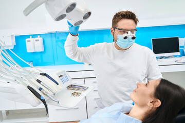 High-qualified dentist in uniform and magnifying dental loupes adjusting light lamp - Powered by Adobe