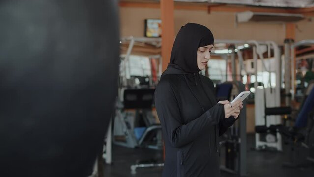 Portrait of a young muslim female athlete in a hijab using a smartphone texting with someone, having a break in an indoor gym in a side view