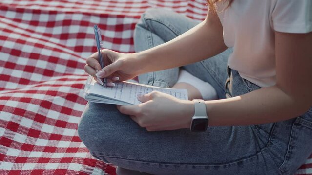 Young girl in casual clothes sitting on the checkered plaid in a city park in summer and taking notes in a notebook close up. Concept of time to rest and meaningfulness in the urban bustle