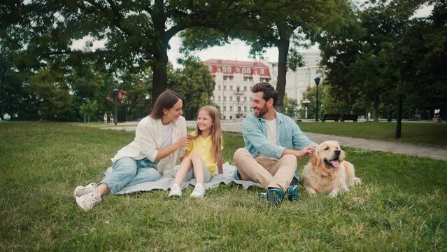 Positive family with dog sits on mat on grass lawn of park