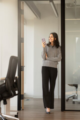 Fototapeta na wymiar Beautiful smiling Indian businesswoman holding mobile phone, looking away in modern office