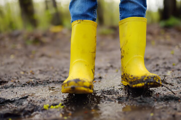 Mischievous preschooler child wearing yellow rain boots jumping in large wet mud puddle. Kid playing and having fun. Outdoors games