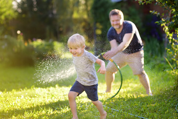 Funny little boy with his father playing with garden hose in sunny backyard. Preschooler child...
