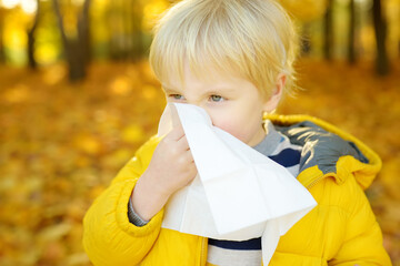 Little boy sneezing and wipes nose with napkin during walking in autumn park