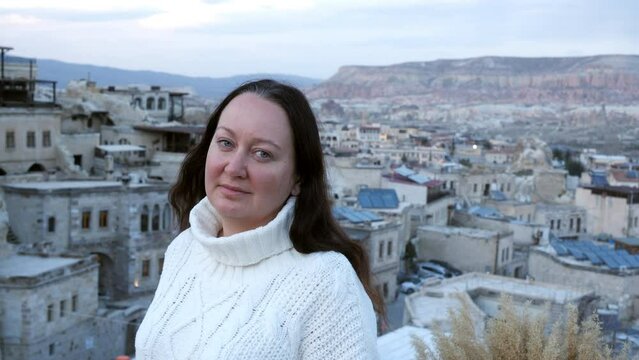 Brunette middle aged woman on terrace balcony looking at camera on ancient rocky town with mountains on background in overcast spring weather. Tourism, travel, vacation, rest and relax, journey.