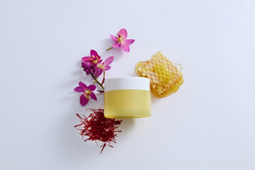 Minimal concept for advertising and branding cosmetic extracted from saffron. A yellow cosmetic jar displayed on white background with stigmas saffron, flowers and beeswax. Mockup for design