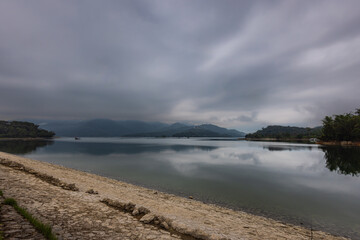 The cloudy sky lends an air of mystique to Sun Moon Lake, Taiwan. Soft light pierces through, casting an ethereal glow on the water. Amidst the mist, a captivating and reflective sight emerges.