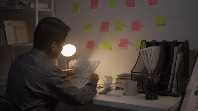 Asian male businessman working with financial documents and desktop computer till late at office, young man working overtime salaryman concept 4k