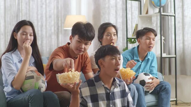 Asian Teenagers Cheering, Eating And Watching Football Game On Tv At Home
