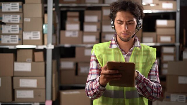 Male worker wearing headset in logistics distribution warehouse using digital tablet for inventory or control