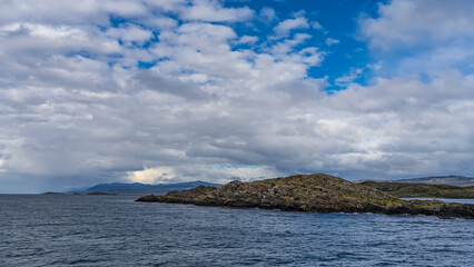 A rocky little island in the Beagle Channel is covered with sparse stunted vegetation. In the distance, against the background of blue sky and clouds - a mountain range. Argentina. 