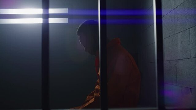 Guilty African American man in orange uniform sits on prison bed and dreams about freedom. Murderer in gloomy correctional facility or detention center. Prisoner serves imprisonment term in jail cell.