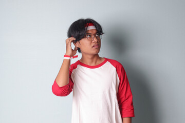 Portrait of attractive Asian man in t-shirt with red and white ribbon on head, standing against gray background, thinking about question with hand on chin