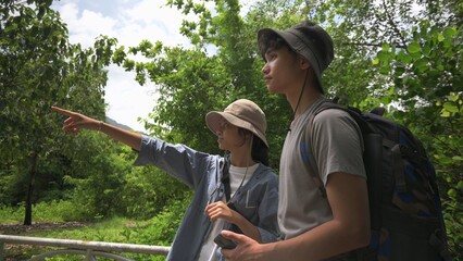 Two Asian young teen backpackers looking for directions while hiking in tropical forest park in summer holiday. Outdoor pursuits. Teenager hobbies and leisure activities. - 628751556