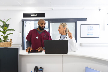 Diverse male and female doctors discussing work and using laptop in reception at hospital