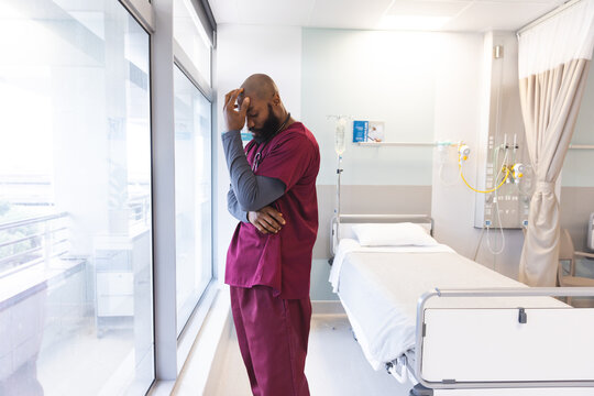 Sad African American Male Doctor Wearing Scrubs Looking Through Window At Hospital