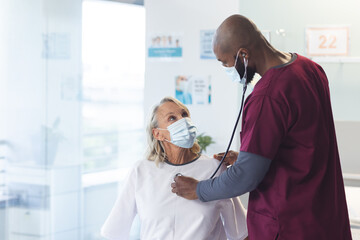 African american doctor examining senior caucasian patient using stethoscope, wearing face masks