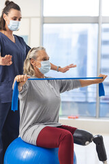 Fototapeta na wymiar Caucasian physiotherapist and senior woman with artificial leg with face masks using exercise band