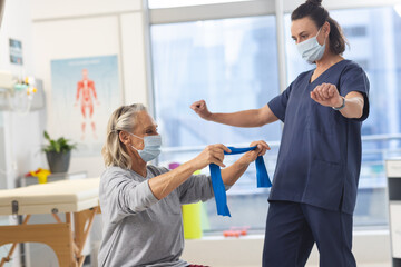 Caucasian physiotherapist and senior woman with face masks using exercise band stretching arms