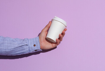 Man's hand in shirt holding white cardboard coffee cup on purple pastel background with shadow