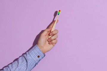 Man's hand in shirt holding rainbow wooden toothbrush on purple pastel background. Lgbt community