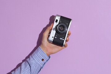 Man's hand in shirt holding retro film camera on purple pastel background with shadow.