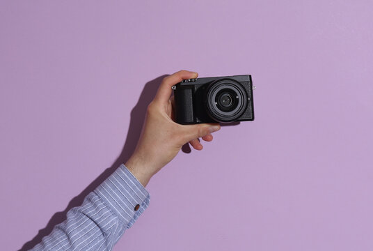 Man's hand in shirt holding modern digital camera on purple pastel background with shadow.