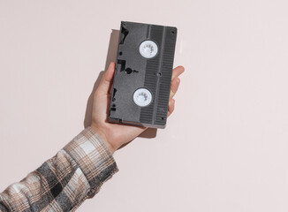 Man's hand in shirt holding vhs video cassette on beige pastel background with shadow.