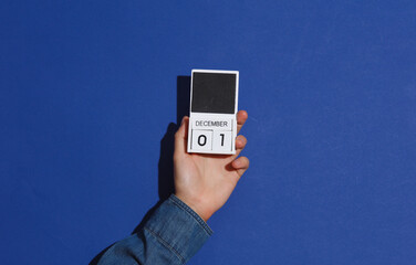 A man's hand in a denim shirt holds a wooden block calendar organizer with the date December 2 on a blue background