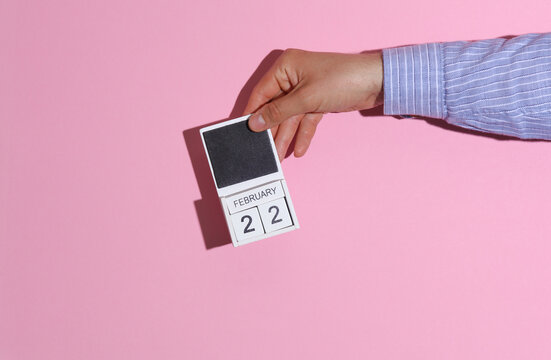 Man's hand in shirt holds wooden block calendar organizer with date February 22 on pink background with shadow
