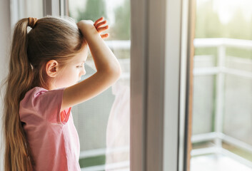 Sad little girl looking through  window at home.