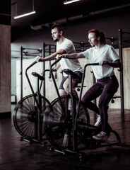 Athletic man and woman doing intense workout together by pedaling air bike in modern gym. Healthy...