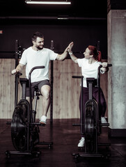 Fototapeta na wymiar Smiling man and woman doing intensive workout together by pedaling air bike and high five in modern gym. Healthy lifestyle