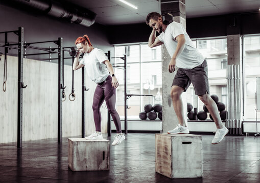 Athletic man and woman practicing lunges together with dumbbell on box in modern gym