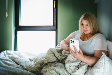 Plus size woman use mobile phone lying in bed. Overweight lady have video call, typing message or browsing social media internet.