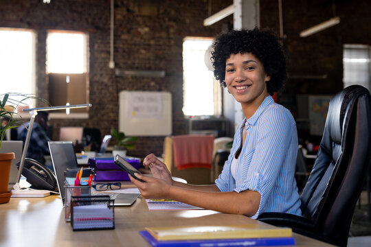 Portrait of happy biracial casual businesswoman sitting at desk using tablet in office