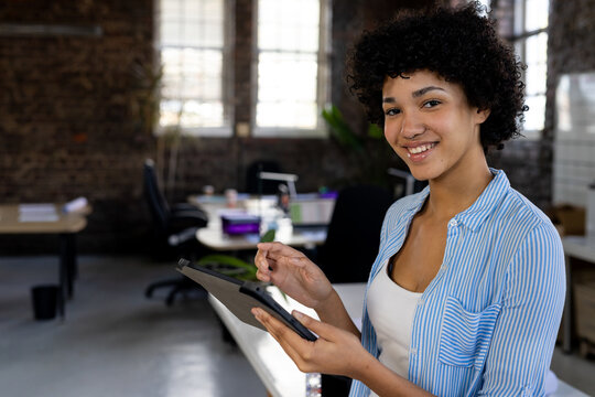 Portrait of happy biracial casual businesswoman standing in office using tablet, copy space