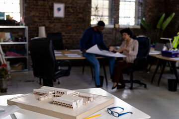 Building model with diverse architects discussing blueprints in background at office, copy space