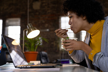 Biracial casual businesswoman using laptop and eating takeaway salad sitting at desk in office