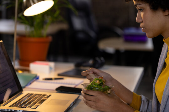 Biracial casual businesswoman using laptop and eating takeaway salad sitting at desk in office