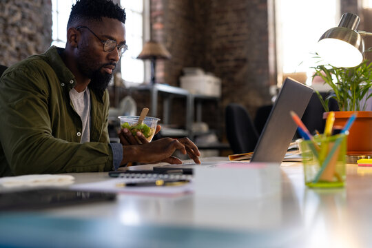 African american casual businessman using laptop and eating takeaway salad sitting at desk in office