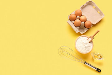 Fototapeta na wymiar Bowl with wheat flour, eggs in package, whisk and oil on yellow background