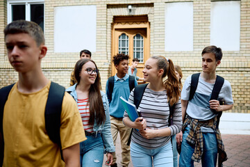 Happy teenage girls talk while getting out of high school building with their classmates.